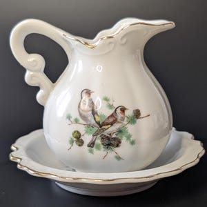 Op de afbeelding: Wit porseleinen roomkan en suikerpot set met gouden rand. De roomkan heeft een vogelontwerp met drie vogels die op een tak zitten.