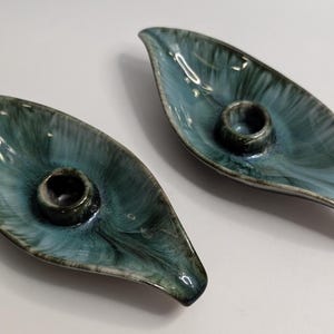Peut inclure: Deux bougeoirs en céramique en forme de feuilles allongées. Chaque pièce présente un vernis brillant avec un motif marbré dans des tons bleu sarcelle profond, bleu et blanc. Une douille circulaire pour bougie est centrée sur chaque base.