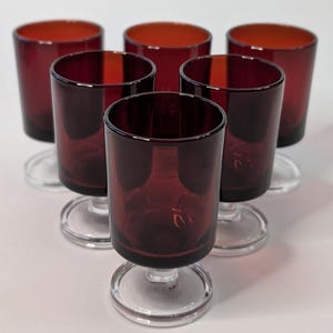 May include: Six vintage wine glasses in a deep burgundy colour. The glasses have a short stem and a clear, circular base. The interior of the glasses has a lighter, reddish-orange colour. The glasses are arranged in a circular pattern.