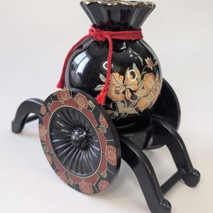 Peut inclure: Un vase décoratif noir avec un motif floral et un cordon rouge, monté sur un chariot noir avec des roues décoratives. Le vase a un bord doré et est orné de fleurs orange et dorées.