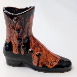 Puede incluir: Un jarrón de cerámica con forma de bota con un esmalte negro y naranja. El jarrón tiene un detalle de botón en el lateral.