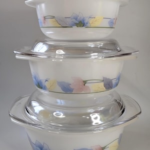 May include: A stack of three white glass bowls with clear glass lids. Each bowl features a horizontal band decorated with a floral pattern in shades of blue, pink, and yellow. The bowls have integrated side handles and a smooth, rounded shape.