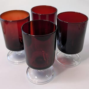 Puede incluir: Cuatro copas de vino tintas de color rojo rubí. Las copas tienen forma cilíndrica con una base transparente y circular. El interior de las copas es de un rojo intenso, con un efecto degradado. Las copas están dispuestas sobre una superficie blanca.