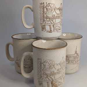 May include: A set of four off-white ceramic mugs with a speckled finish and brown rim. Each mug features a brown line-art illustration of a coastal village scene with buildings, boats, and a church steeple, all with a matching side handle.