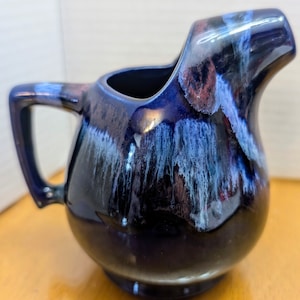 Peut inclure: Une crémier en céramique bleue avec un glaçage blanc et violet. La crémier a un corps arrondi et un bec.