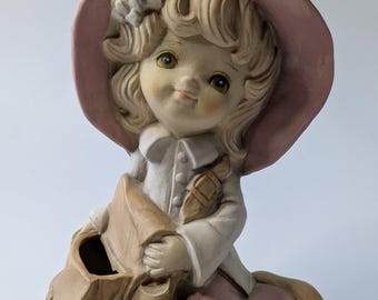 Könnte beinhalten: Eine Keramikfigur eines Mädchens, das einen rosa Hut und ein rosa Kleid trägt und eine braune Tasche hält. Die Tasche hat eine Öffnung zum Pflanzen.