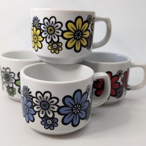 Op de afbeelding: Vier witte keramische mokken met kleurrijke bloemmotieven. De bloemen zijn geel, blauw, rood en groen, omlijnd met zwart. Elke mok heeft een klein handvat. Geschikt voor koffie of thee.