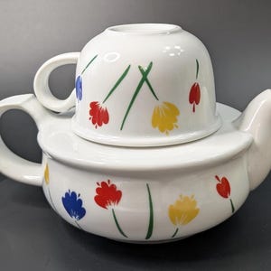 May include: White ceramic teapot and teacup set. The teapot has a rounded shape with a handle and spout. The teacup sits on top of the teapot. Both pieces are decorated with colourful tulip designs in red, yellow, and blue.