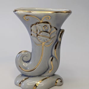 Peut inclure: Un vase en céramique bleu clair avec une bordure dorée et un motif de rose en relief. Le vase a la forme d'une corne.
