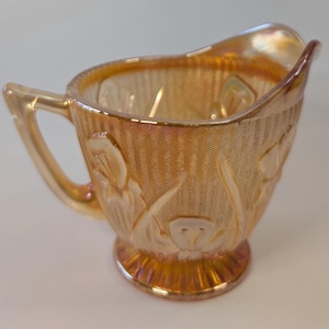 Peut inclure: Un pot à lait en verre carnaval couleur souci avec une finition irisée et un motif floral en relief représentant des iris. Le récipient présente un fond texturé à chevrons, un bec verseur évasé, une anse courbée et une base piédouche.