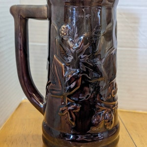 Puede incluir: Una jarra de cerveza de cerámica marrón con un diseño en relieve de la cara de un hombre y un árbol. La jarra tiene un asa y mide aproximadamente 25 cm de alto.