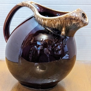 Peut inclure: Un pichet en céramique émaillée brun et crème avec un corps arrondi et un bec large. Le pichet a une finition brillante et un motif unique de glaçure brun et crème.