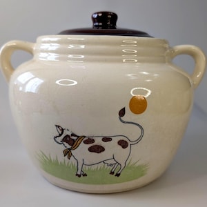 May include: Cream ceramic cookie jar with a brown lid and handles. The jar features a cartoon cow illustration with brown spots, a yellow scarf, and a green grass base. The lid has a dark brown knob.