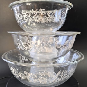 May include: A set of three clear glass mixing bowls stacked vertically. Each bowl features a white floral pattern consisting of daisies and leafy stems encircling the exterior. The bowls have a smooth, rounded rim and a transparent, durable glass construction.