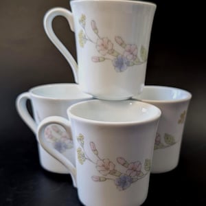 May include: A set of four white ceramic mugs stacked together. Each mug features a delicate floral pattern with small pink, blue, and yellow flowers along with green leaves. The mugs have a smooth, cylindrical shape with a single curved handle on the side.