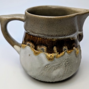 Peut inclure: Un petit pot à crème en céramique brun et blanc avec une surface texturée et une bande brune autour du milieu.