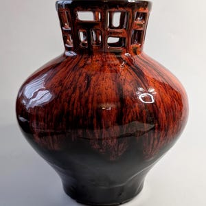 Puede incluir: Un jarrón decorativo de cerámica con acabado brillante. El jarrón presenta una combinación de colores marrón oscuro y negro con un diseño calado único en la parte superior. El cuerpo del jarrón tiene forma redondeada.