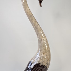 Może przedstawiać: Wysoka ceramiczna figurka łabędzia z zakrzywioną szyją i ciemnobrązową głową i podstawą. Korpus to mieszanka jasnobrązowego i białego, z błyszczącym wykończeniem. Łabędź to przedmiot dekoracyjny.
