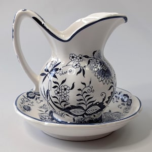 Può includere: Un'anfora in ceramica bianca con un motivo floreale blu e un sottopiatto abbinato. L'anfora ha un'ampia apertura e un manico.