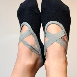 Könnte beinhalten: Ein Paar schwarze Socken mit niedrigem Schnitt und hellgrauen elastischen Riemen, die sich über den Fußrücken kreuzen. Die Socken haben eine abgerundete Zehenpartie und einen tiefen Ausschnitt, der den Fußrücken für einen sicheren Halt frei lässt.