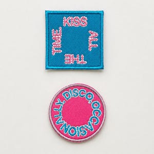 Può includere: Due toppe ricamate su sfondo bianco. Una è quadrata, color verde acqua con testo rosa che recita "Kiss All The Time". L'altra è un cerchio rosa con le parole "Disco Occasionally" in verde acqua.