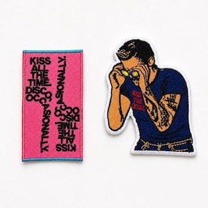 May include: Two embroidered patches. One is a rectangular pink patch with a blue border featuring the text KISS ALL THE TIME. KISS OCCASIONALLY. The other is a shaped patch of a person in a blue t-shirt with yellow glasses and tattoos.