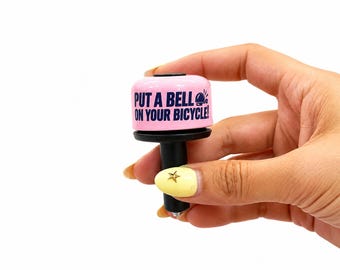 May include: A pink bicycle bell with a black base and black text that reads "PUT A BELL ON YOUR BICYCLE!" accompanied by a small icon of a bell. The bell features a cylindrical shape and a smooth finish, designed for attachment to handlebars.