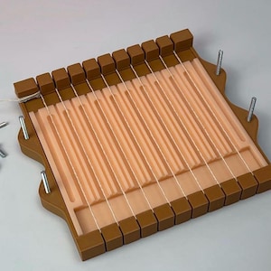 May include: A brown and peach-colored weaving loom with white threads strung across. The loom has a rectangular frame with a series of vertical slots and a row of square blocks along the top and bottom edges. Metal pins are visible on the sides.
