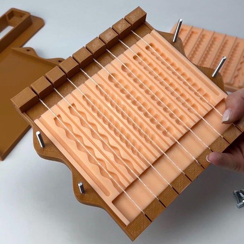 Puede incluir: Un telar de madera marr&oacute;n con un interior color melocot&oacute;n y hilos blancos. El telar presenta varios patrones texturizados. Piezas marrones adicionales son visibles en el fondo.