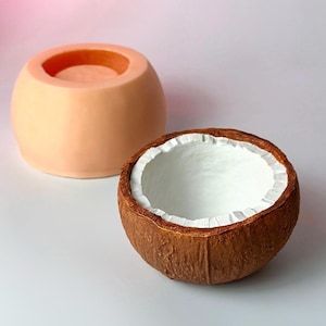 May include: A halved coconut with a white interior and a brown, textured exterior sits next to a peach-colored, round mold. The mold has a circular indentation on top. The items are on a white surface.