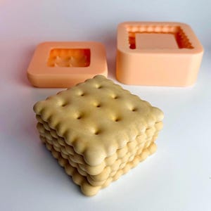 Puede incluir: Una pila de galletas cuadradas de color marrón claro con bordes redondeados y pequeños agujeros. Dos moldes de silicona de color melocotón están en el fondo, uno cuadrado y otro rectangular, diseñados para crear la forma de la galleta.