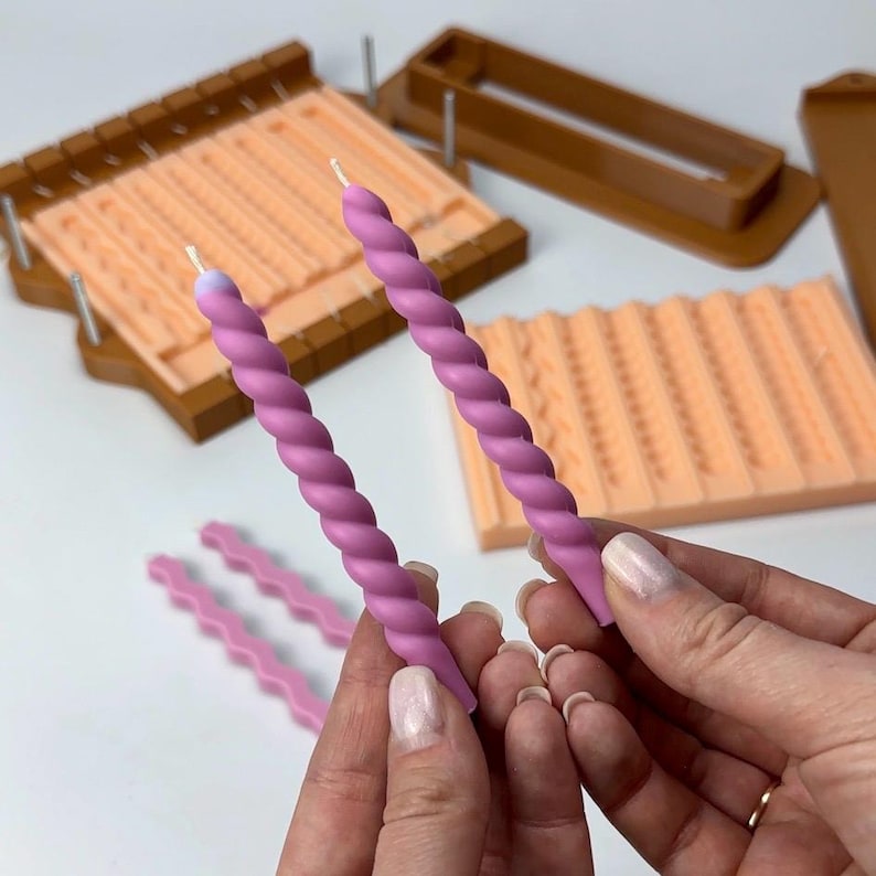 Puede incluir: Dos velas retorcidas de color lavanda se sostienen frente a moldes para velas. Los moldes son marrones y tienen una superficie texturizada. Tambi&eacute;n se ven varias otras velas de lavanda. La imagen muestra suministros para hacer velas.