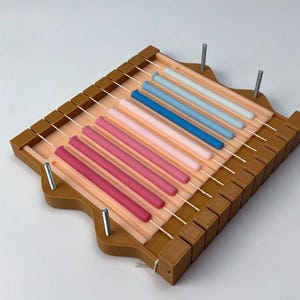 Puede incluir: Un molde de madera marrón para hacer velas con una serie de cuerdas paralelas. Contiene velas cilíndricas de colores en tonos rosa, azul y blanco. Los pasadores metálicos aseguran la estructura.