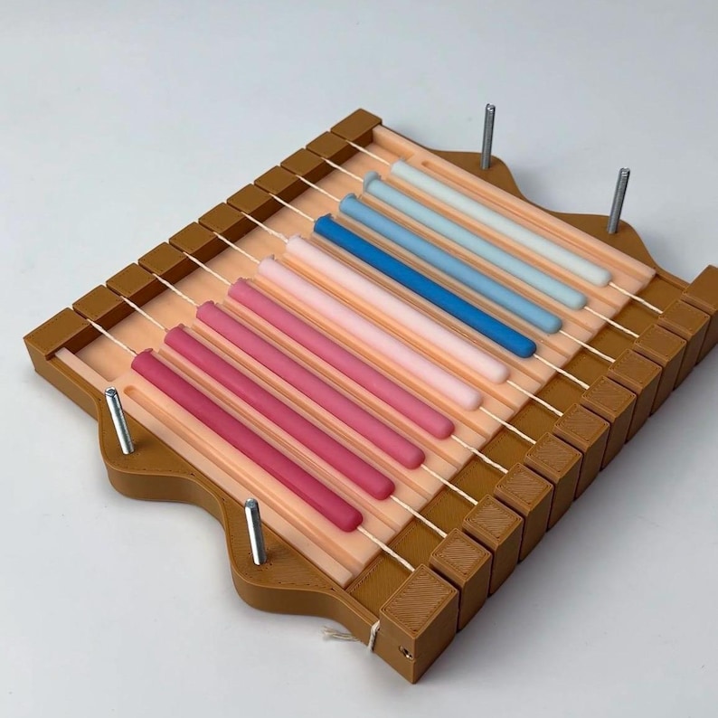 May include: A brown wooden candle making mold with a series of parallel strings. The mold holds a variety of colorful, cylindrical candles in shades of pink, blue, and white. Metal pins secure the mold's structure.