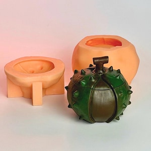 Può includere: Un contenitore a forma di castagna verde scuro e marrone con un gambo, accompagnato da due stampi in silicone arancione chiaro. La castagna presenta un esterno spinoso e testurizzato con una cucitura centrale, mentre gli stampi mostrano lo spazio vuoto.