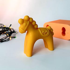 May include: A yellow toy horse with a decorative saddle and mane. The horse is made of a solid material and has a rounded shape. An orange mold is in the background. The horse is a toy.