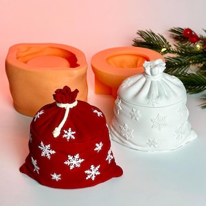 Pode incluir: Um recipiente vermelho com tema natalino em forma de saco, adornado com desenhos de flocos de neve brancos. Um recipiente branco com desenhos de flocos de neve também está presente. Dois moldes laranja estão no fundo. Decoração festiva de feriado.