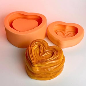 Op de afbeelding: Een set oranje siliconen hartvormige mallen, samen met een afgewerkt goudbruin hartvormig object. De mallen zijn ontworpen met gelaagde hartvormen. Het afgewerkte object heeft een textuur.