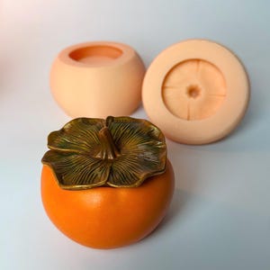 Op de afbeelding: Een oranje, op een kaki lijkende container met een gedetailleerd bronzen deksel. Twee extra perzik-kleurige, ronde containers staan op de achtergrond. Het deksel heeft een gedetailleerd bladerenpatroon. De container is waarschijnlijk voor decoratieve of opslagdoeleinden.