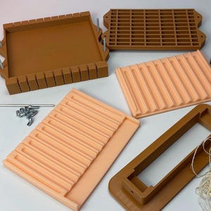 May include: A collection of brown and peach-colored soap molds and accessories. The molds include rectangular trays with dividers, a grid-patterned mold, and a frame. Metal rods and hardware are also present, suggesting a DIY soap-making setup.