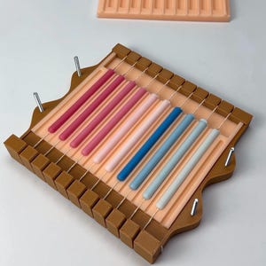 May include: A candle-making kit featuring a rectangular, light brown frame with a peach-colored interior. It holds several colorful, cylindrical candles in shades of pink, blue, and white. The kit includes a second, similar frame in the background.