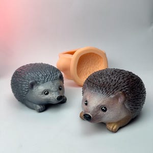 Könnte beinhalten: Zwei detaillierte Igel-Figuren, eine graue und eine braune, befinden sich in der Nähe eines pfirsichfarbenen, oben offenen Behälters. Die Igel haben kleine, schwarze Augen und sind vor einem schlichten, weißen Hintergrund platziert.