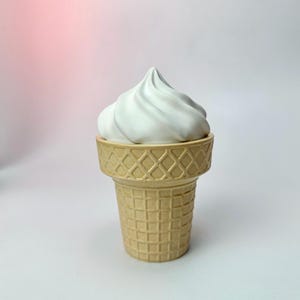 Op de afbeelding: Een keramische ijshoorntje met een witte werveling van softijs. De kegel heeft een lichtbruine kleur met een wafelpatroon. De achtergrond is een verloop van roze en wit.
