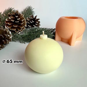 Peut inclure: Une bougie sphérique jaune pâle avec une mèche, ressemblant à une boule de Noël. La bougie mesure 65 mm de diamètre. Un pot de fleurs sphérique pêche avec une base unique est également visible, ainsi que des pommes de pin et des branches de conifères.