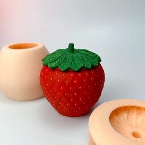 Könnte beinhalten: Ein roter und grüner Keramikbehälter in Form einer Erdbeere mit Deckel. Der Behälter ist so gestaltet, dass er wie eine Erdbeere mit grünem, blattreichem Oberteil aussieht. Der Behälter steht auf einer weißen Oberfläche.