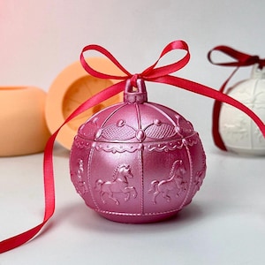 Könnte beinhalten: Eine rosa Schmuckschatulle im Karussell-Stil mit einer roten Schleife. Die runde Schatulle zeigt geprägte Karussellpferde und dekorative Details. Weitere dekorative Gegenstände befinden sich im Hintergrund.