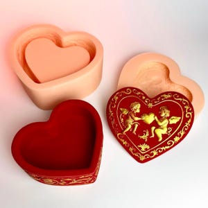 Puede incluir: Una caja de baratijas roja en forma de corazón con detalles dorados y un borde decorativo. También son visibles dos moldes en forma de corazón, uno rojo y otro melocotón. También hay un objeto rojo en forma de corazón con querubines dorados.