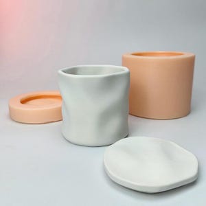 Puede incluir: Una taza de cerámica blanca con un aspecto texturizado y arrugado, un platillo blanco a juego y dos moldes de silicona color melocotón. Los moldes son una tapa pequeña y redonda y un recipiente cilíndrico. Los artículos están sobre una superficie blanca.