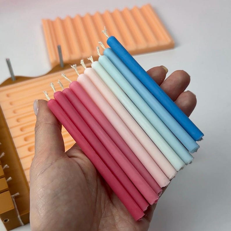 May include: A hand holds a collection of colorful taper candles in shades of pink, red, blue, and white. The candles are arranged in a gradient, with a light orange candle mold in the background.