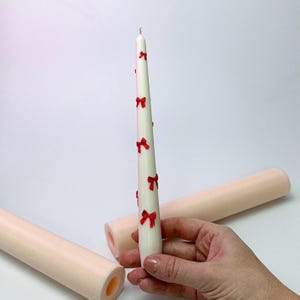 Könnte beinhalten: Eine weiße konische Kerze, verziert mit roten Schleifen. Die Kerze wird von einer Hand gehalten, mit zwei unbeleuchteten, hellrosa Kerzenformen im Hintergrund. Die Kerze ist ein dekorativer Gegenstand.